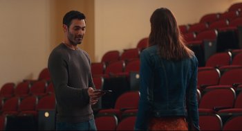 Movie still from “Can You Keep a Secret?” (2019), directed by Elise Durán – A man and a woman standing next to each other in front of red chairs; Medium shot, Over the shoulder angle