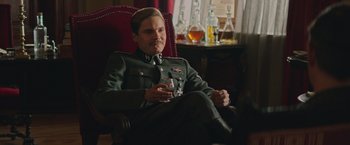 Movie still from “The Zookeeper's Wife” (2017), directed by Niki Caro – A man in a uniform sitting in a chair holding a drink; Medium shot, Over the shoulder angle