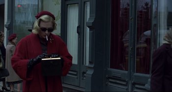 Movie still from “Carol” (2015), directed by Todd Haynes – A woman in a red coat smoking a cigarette and looking at a book; Medium shot, Over the shoulder angle