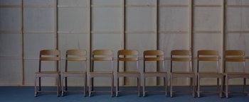 Movie still from “Casting JonBenet” (2017), directed by Kitty Green – A row of wooden chairs sitting in front of a wall; Wide shot, High angle