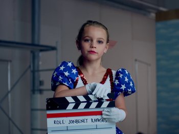 Movie still from “Casting JonBenet” (2017), directed by Kitty Green – A young girl holding a clapperboard in front of her face; Medium shot, Low angle