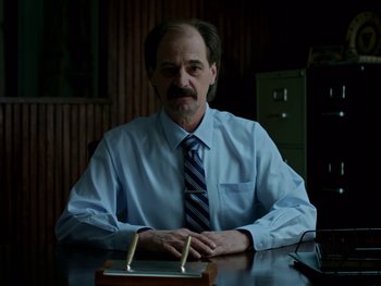Movie still from “Casting JonBenet” (2017), directed by Kitty Green – A man sitting at a desk in front of an open book; Medium shot, Low angle