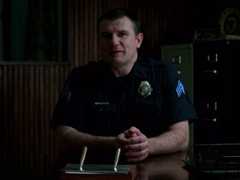 Movie still from “Casting JonBenet” (2017), directed by Kitty Green – A police officer sitting at a table in front of a computer; Medium shot, Low angle