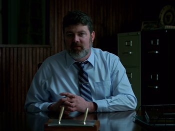 Movie still from “Casting JonBenet” (2017), directed by Kitty Green – A man sitting at a table in front of an open filing cabinet; Medium shot, Over the shoulder angle