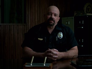 Movie still from “Casting JonBenet” (2017), directed by Kitty Green – A police officer sitting at a table with his arms crossed; Medium shot, Over the shoulder angle