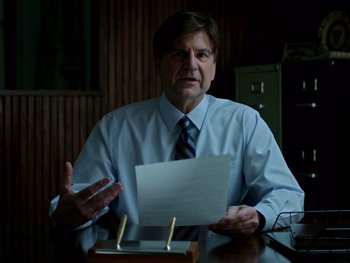 Movie still from “Casting JonBenet” (2017), directed by Kitty Green – A man sitting at a desk holding a piece of paper; Medium shot, Over the shoulder angle