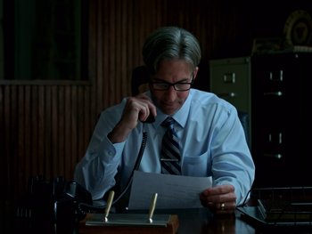 Movie still from “Casting JonBenet” (2017), directed by Kitty Green – A man sitting at a desk holding a phone to his ear; Medium shot, Low angle