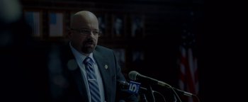 Movie still from “Casting JonBenet” (2017), directed by Kitty Green – A man in a suit and tie speaking into microphones; Close Up shot, Over the shoulder angle