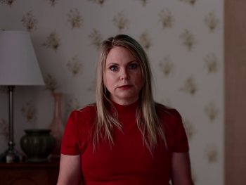 Movie still from “Casting JonBenet” (2017), directed by Kitty Green – A woman wearing a red shirt standing next to a lamp; Medium shot, Low angle