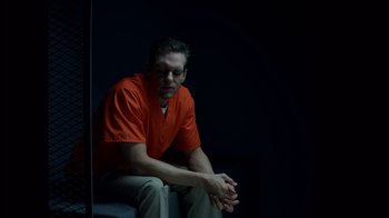 Movie still from “Casting JonBenet” (2017), directed by Kitty Green – A man in an orange shirt sitting in a dark room; Medium shot, High angle