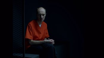 Movie still from “Casting JonBenet” (2017), directed by Kitty Green – A man in an orange shirt sitting in a dark room; Medium shot, High angle