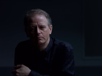 Movie still from “Casting JonBenet” (2017), directed by Kitty Green – A man in a black shirt is sitting in a dark room; Close Up shot, Low angle