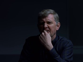 Movie still from “Casting JonBenet” (2017), directed by Kitty Green – An older man is sitting in a dark room; Close Up shot, Over the shoulder angle