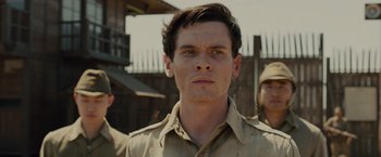 Movie still from “Unbroken” (2014), directed by Angelina Jolie – A young man in a tan shirt is standing in front of a fence; Close Up shot, Low angle