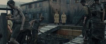 Movie still from “Unbroken” (2014), directed by Angelina Jolie – Two men in uniforms stand in front of a building while a man in a gray t - shirt looks on; Wide shot, High angle