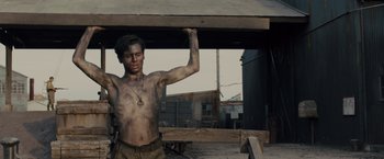 Movie still from “Unbroken” (2014), directed by Angelina Jolie – A shirtless man standing next to a wooden structure; Medium shot, Low angle