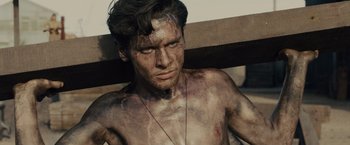 Movie still from “Unbroken” (2014), directed by Angelina Jolie – A shirtless man with brown hair and a necklace; Close Up shot, Low angle