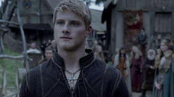 Movie still from “Vikings” (2013), created by Michael Hirst – A man with a cross necklace standing in front of a crowd of people; Close Up shot, Over the shoulder angle