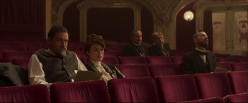 Movie still from “Colette” (2018), directed by Wash Westmoreland – A woman sitting in a red chair in front of two other people; Medium shot, High angle