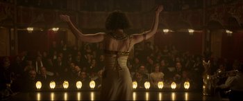Movie still from “Colette” (2018), directed by Wash Westmoreland – A woman standing in front of an audience in front of lights; Medium shot, Over the shoulder angle
