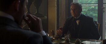 Movie still from “Colette” (2018), directed by Wash Westmoreland – A man sitting at a table with a cigarette in his hand; Medium shot, Over the shoulder angle