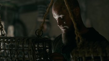 Movie still from “Vikings” (2013), created by Michael Hirst – A bearded man holding a bird cage in a dark room; Close Up shot, Low angle