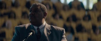 Movie still from “Come Sunday” (2018), directed by Joshua Marston – A man wearing a suit and tie standing in front of a microphone in front of a crowd; Close Up shot, Low angle