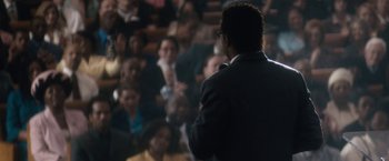 Movie still from “Come Sunday” (2018), directed by Joshua Marston – A man standing in front of a crowd of people; Medium shot, Over the shoulder angle