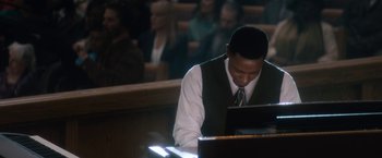 Movie still from “Come Sunday” (2018), directed by Joshua Marston – A man sitting at a piano in front of a crowd of onlookers; Medium shot, High angle