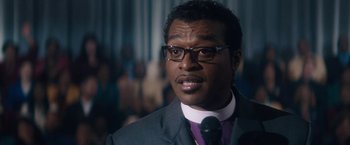 Movie still from “Come Sunday” (2018), directed by Joshua Marston – A person wearing a suit and glasses; Close Up shot, Over the shoulder angle