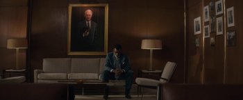 Movie still from “Come Sunday” (2018), directed by Joshua Marston – A man in a suit sitting on a couch next to a painting; Wide shot, Over the shoulder angle
