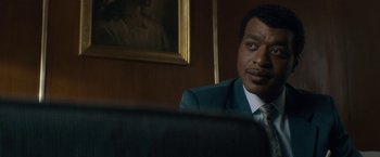 Movie still from “Come Sunday” (2018), directed by Joshua Marston – A man sitting in a chair in front of a painting; Close Up shot, Over the shoulder angle