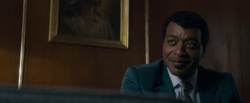 Movie still from “Come Sunday” (2018), directed by Joshua Marston – A man sitting in front of a painting in a room; Close Up shot, Over the shoulder angle