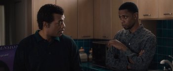 Movie still from “Come Sunday” (2018), directed by Joshua Marston – Two men are standing in a kitchen talking to each other; Medium shot, Over the shoulder angle