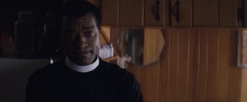 Movie still from “Come Sunday” (2018), directed by Joshua Marston – A man wearing a priest's outfit in a kitchen; Close Up shot, Over the shoulder angle