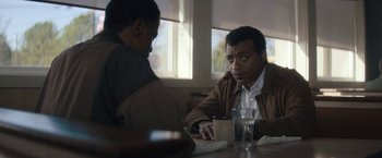 Movie still from “Come Sunday” (2018), directed by Joshua Marston – Two men sitting at a table with a cup of coffee; Medium shot, Over the shoulder angle