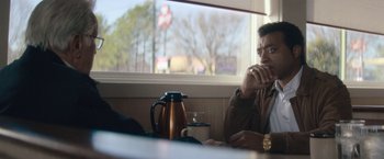 Movie still from “Come Sunday” (2018), directed by Joshua Marston – A man sitting at a table in front of a coffee pot; Medium shot, Over the shoulder angle