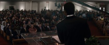 Movie still from “Come Sunday” (2018), directed by Joshua Marston – A man is speaking at a public speaking event; Wide shot, Over the shoulder angle
