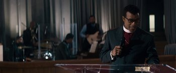 Movie still from “Come Sunday” (2018), directed by Joshua Marston – A man is holding a microphone in front of a microphone stand; Medium shot, Over the shoulder angle