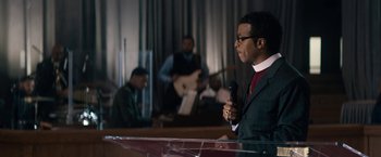 Movie still from “Come Sunday” (2018), directed by Joshua Marston – A man is holding a microphone in front of other people; Medium shot, Over the shoulder angle