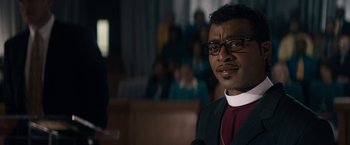 Movie still from “Come Sunday” (2018), directed by Joshua Marston – A man wearing glasses and a priest's outfit; Close Up shot, Low angle