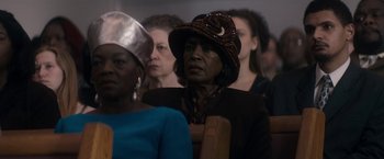 Movie still from “Come Sunday” (2018), directed by Joshua Marston – An older woman wearing a brown hat in front of a group of other women; Close Up shot, Low angle