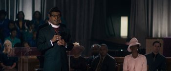 Movie still from “Come Sunday” (2018), directed by Joshua Marston – A man standing in front of a microphone in front of other people; Medium shot, Over the shoulder angle