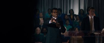 Movie still from “Come Sunday” (2018), directed by Joshua Marston – A man in a black suit and a red tie; Medium shot, Over the shoulder angle