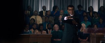 Movie still from “Come Sunday” (2018), directed by Joshua Marston – A man giving a speech in front of an audience; Medium shot, Over the shoulder angle
