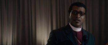 Movie still from “Come Sunday” (2018), directed by Joshua Marston – A man wearing a suit and tie in front of a curtain; Close Up shot, Low angle