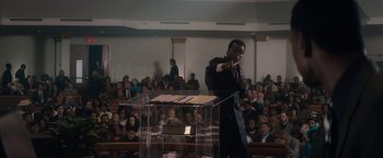 Movie still from “Come Sunday” (2018), directed by Joshua Marston – A man is giving a speech in front of an audience; Medium shot, High angle