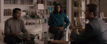 Movie still from “Come Sunday” (2018), directed by Joshua Marston – A woman sitting on a chair in a living room; Medium shot, Over the shoulder angle