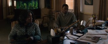 Movie still from “Come Sunday” (2018), directed by Joshua Marston – A man and woman sitting at a table in front of a television; Medium shot, Over the shoulder angle