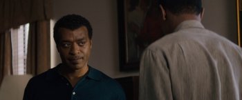 Movie still from “Come Sunday” (2018), directed by Joshua Marston – A man standing next to another man in front of a painting; Close Up shot, Over the shoulder angle
